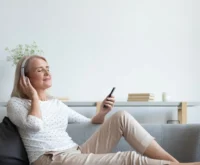 Calm mature woman relax on cozy couch at home wear earphones enjoy music using modern cellphone, modern senior female gadget user in headphones listen to favorite track good sound on smartphone