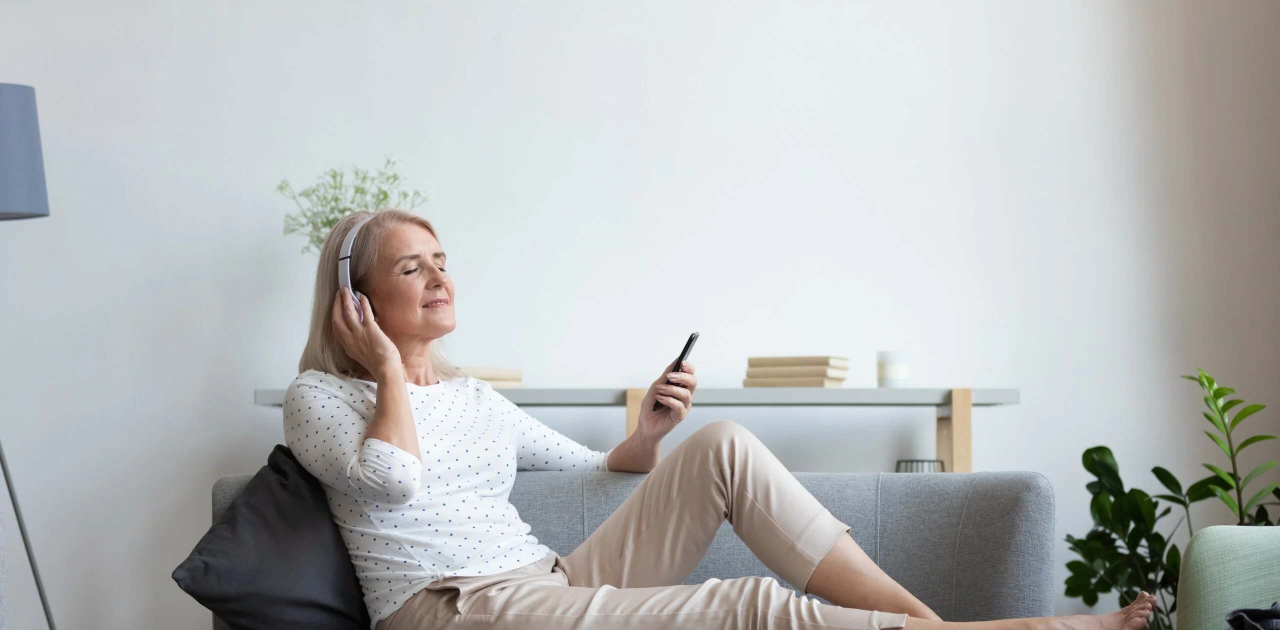 Calm mature woman relax on cozy couch at home wear earphones enjoy music using modern cellphone, modern senior female gadget user in headphones listen to favorite track good sound on smartphone