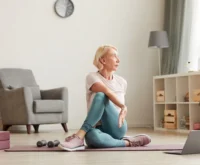 Senior woman sitting on the floor on exercise mat in front of laptop and training at home