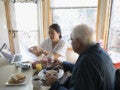 Home Caregiver Explaining Paperwork To Senior Man Eating Breakfast At Dining Table