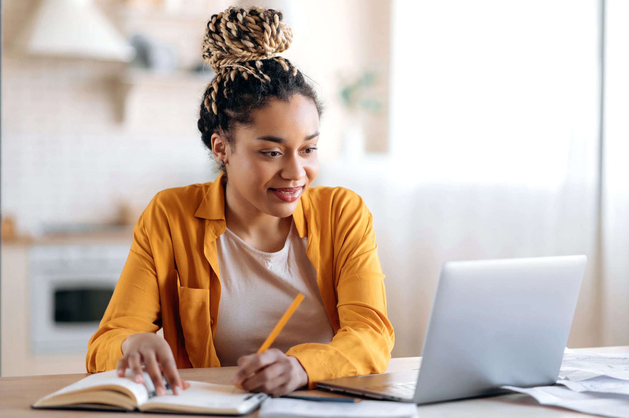 Focused stylish african american female student with afro dreadlocks, studying remotely from home, using a laptop, taking notes on notepad during online lesson, e-learning concept, smiling stock photo