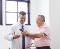 A healthcare professional showing a mobile phone screen to an elderly male patient, illustrating the use of digital tools to support medication adherence and patient education