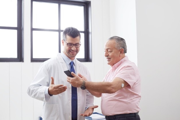 A healthcare professional showing a mobile phone screen to an elderly male patient, illustrating the use of digital tools to support medication adherence and patient education