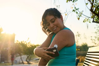 Person scanning continuous glucose monitor (CGM) with smartphone in park at sunset, promoting diabetes self-management and treatment adherence.