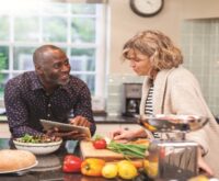 A man and a woman around a kitchen table piled with fresh produce and bread. Man holding a tablet talking to a woman who is cutting vegetables on the counter.