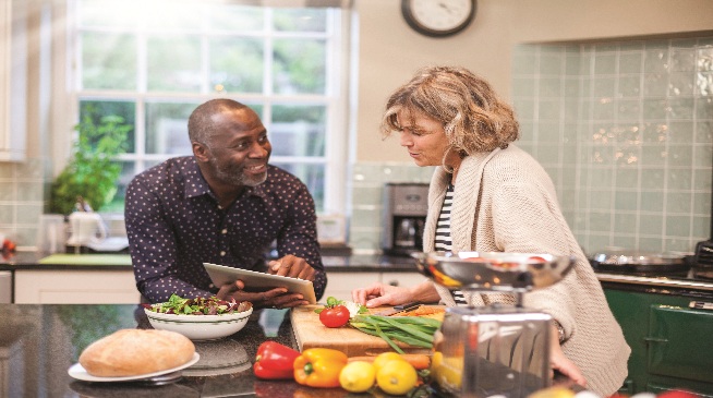 A man and a woman around a kitchen table piled with fresh produce and bread. Man holding a tablet talking to a woman who is cutting vegetables on the counter.