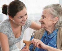 An older woman, sitting on a chair with a walking stick in hand, is talking to a younger woman who is smiling and listening to her.