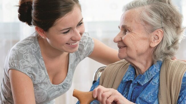 An older woman, sitting on a chair with a walking stick in hand, is talking to a younger woman who is smiling and listening to her.