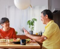 A man and a woman sit at a dining table, engaged in conversation, while the man holds a mobile phone in his hand.