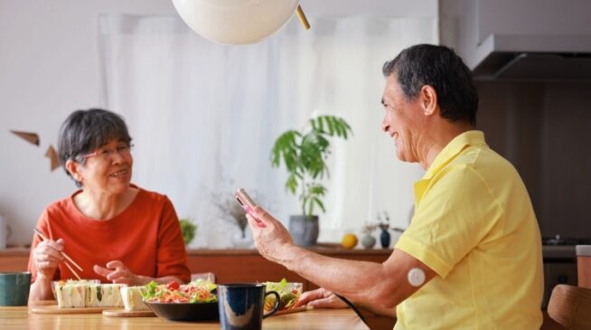 A man and a woman sit at a dining table, engaged in conversation, while the man holds a mobile phone in his hand.
